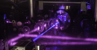 JILA's three-dimensional (3-D) quantum gas atomic clock consists of a grid of light formed by three pairs of laser beams. Multiple lasers of various colors are used to cool the atoms, trap them in a grid of light, and probe them for clock operation. A blue laser beam excites a cube-shaped cloud of strontium atoms. Strontium atoms fluorescence strongly when excited with blue light, as seen in the upper right corner behind the vacuum window.